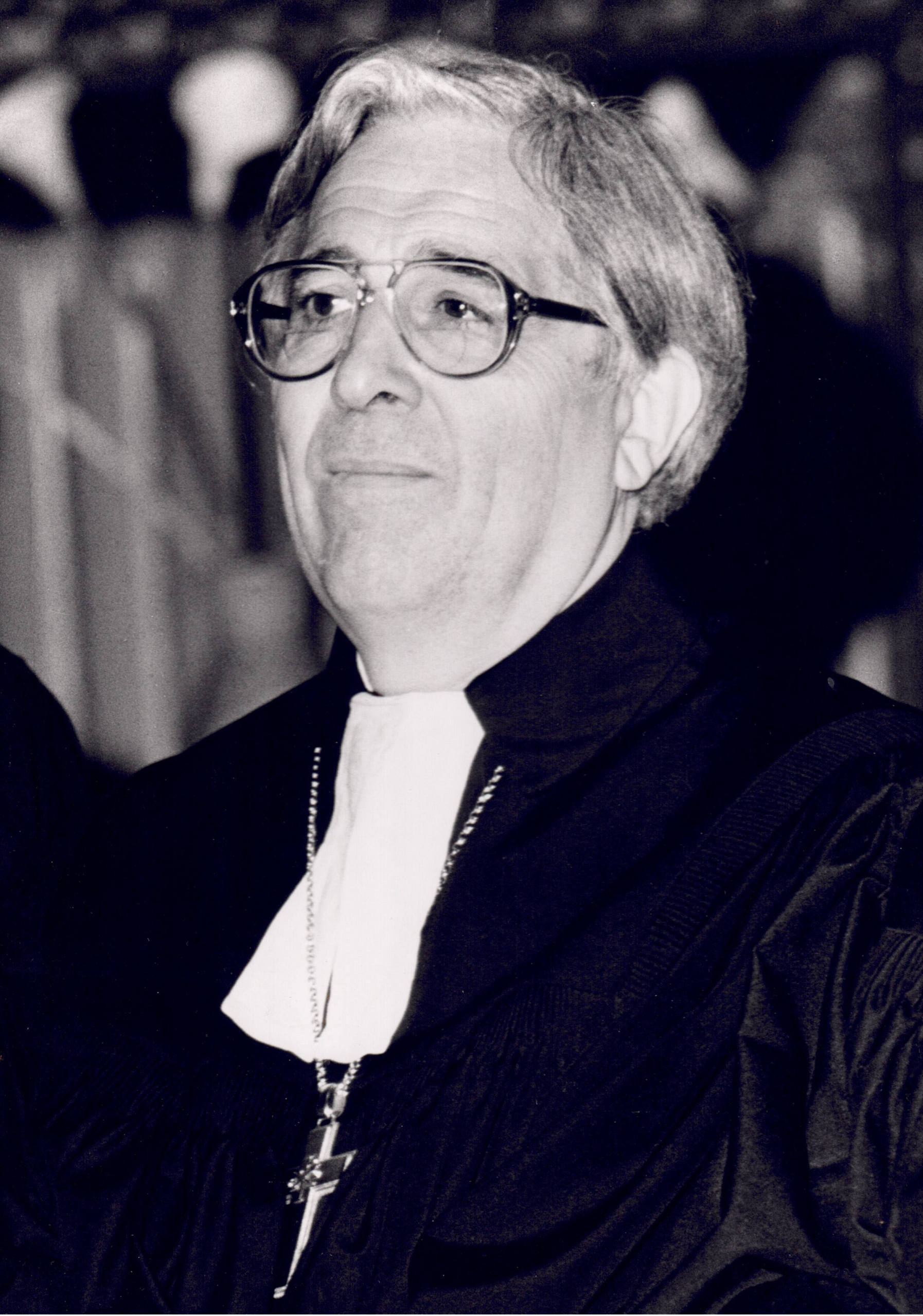 Eine ältere Person mit Brille, die ein religiöses Gewand und eine Kreuzkette trägt, blickt leicht nach oben. Der Hintergrund ist unscharf und in schwarz-weiß gehalten.